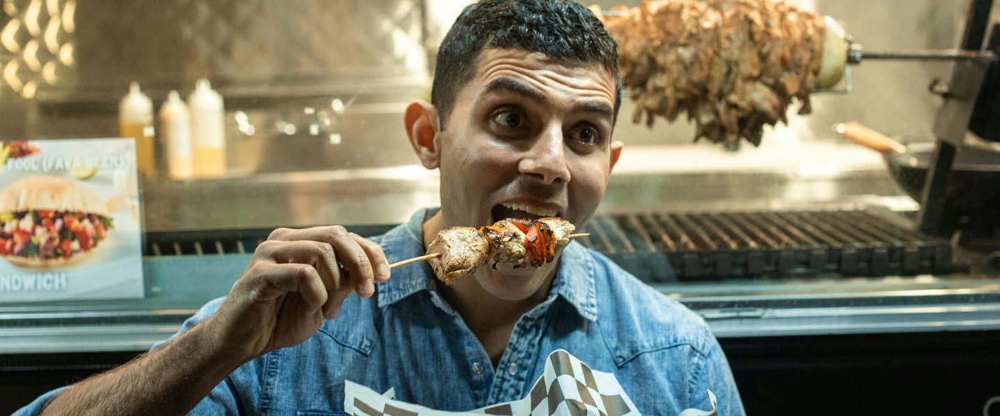 Young man eating a meat kebab skewer with a surprised and happy expression. He is stood in front of a kebab grill with a large joint of meat cooking in the background.