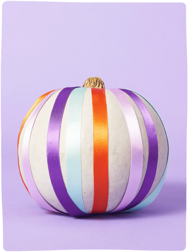Pale green pumpkin decorated with colourful ribbons from top to bottom sat against a light purple backdrop.