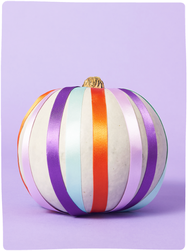 Pale green pumpkin decorated with colourful ribbons from top to bottom sat against a light purple backdrop.